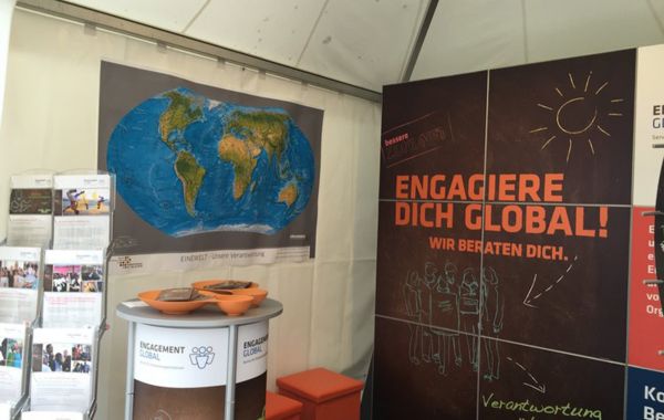 Informationsstand in einem Zelt mit Banner „Engagiere dich global! Wir beraten dich. Verantwortung übernehmen“. Links eine Weltkarte mit dem Titel „Unsere Veranstaltungen“ und ein Tisch mit Broschüren.
