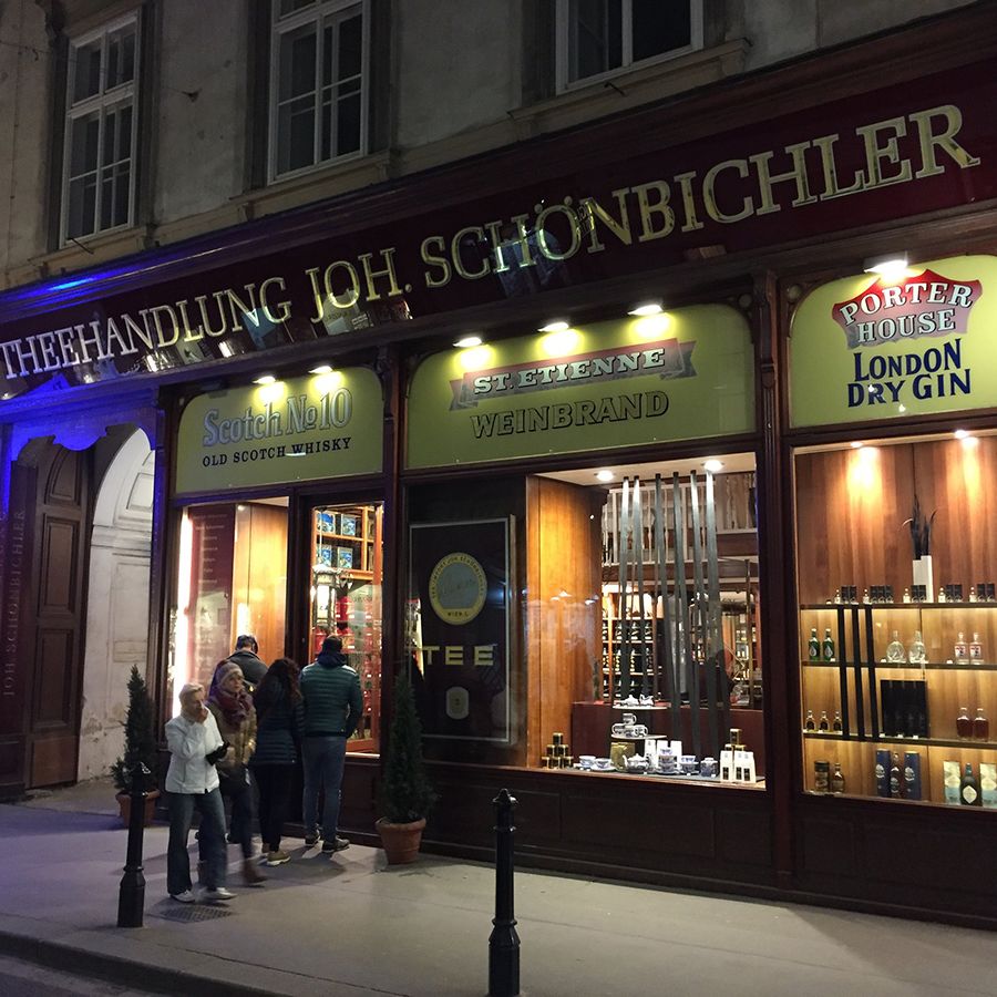 Beleuchtetes Schaufenster eines Spirituosengeschäfts bei Nacht. Schild „THEEHANDLUNG JOH. SCHÖNBICHLER“, Flaschen und Marken sichtbar. Mehrere Personen davor.