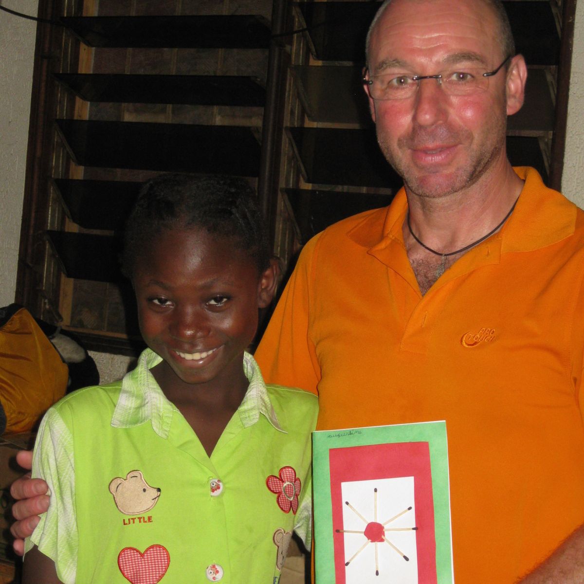 Ein Kind in grünem Kleid mit Bärchenmotiv steht neben einem Erwachsenen mit orangefarbenem Shirt und Brille. Der Erwachsene hält eine Karte mit geometrischem Muster.