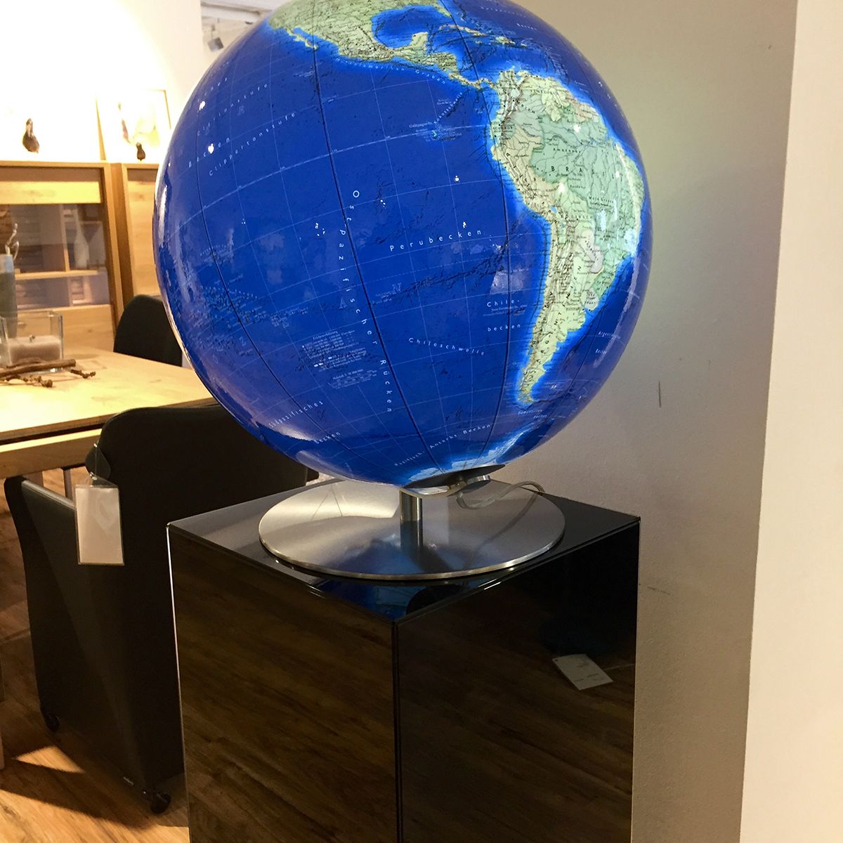 Ein beleuchteter Globus auf einem dunklen Sockel zeigt Nord- und Südamerika. Im Hintergrund sind Möbelstücke wie Tisch, Stühle und Sofa in einem Innenraum sichtbar.