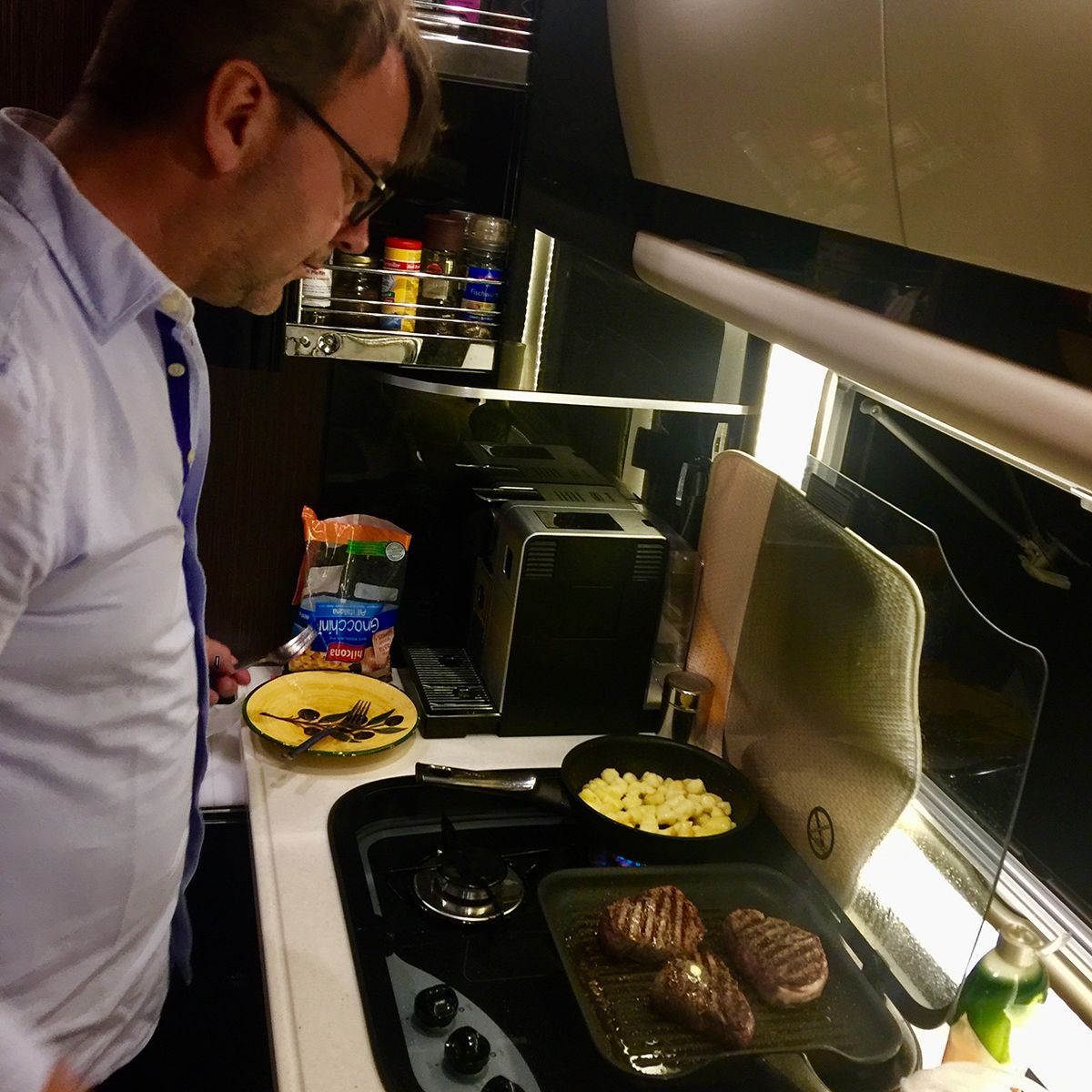 Ein Mann mit Brille kocht in einer modernen Küche. Auf dem Herd braten Fleischstücke und Kartoffelwürfel. Auf der Arbeitsfläche stehen ein Teller mit Pilzen, ein Butterstück und Lebensmittelverpackungen.