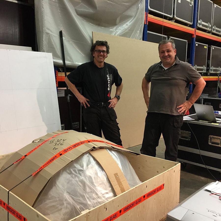 Zwei Personen neben einer Holzkiste mit verpacktem Kuppelobjekt, beschriftet mit „FRAGILE“. Umgebung mit Regalen, Transportkoffern und Laptop.