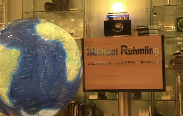 nnenraum mit großem Globus, Holzschild mit der Aufschrift „Michael Ruhmling – OBJEKTE · IDEEN · KUNST“ und beleuchtetem Spiralmotiv darüber. Im Hintergrund Glasregale mit Büsten, Büchern und dekorativen Objekten.