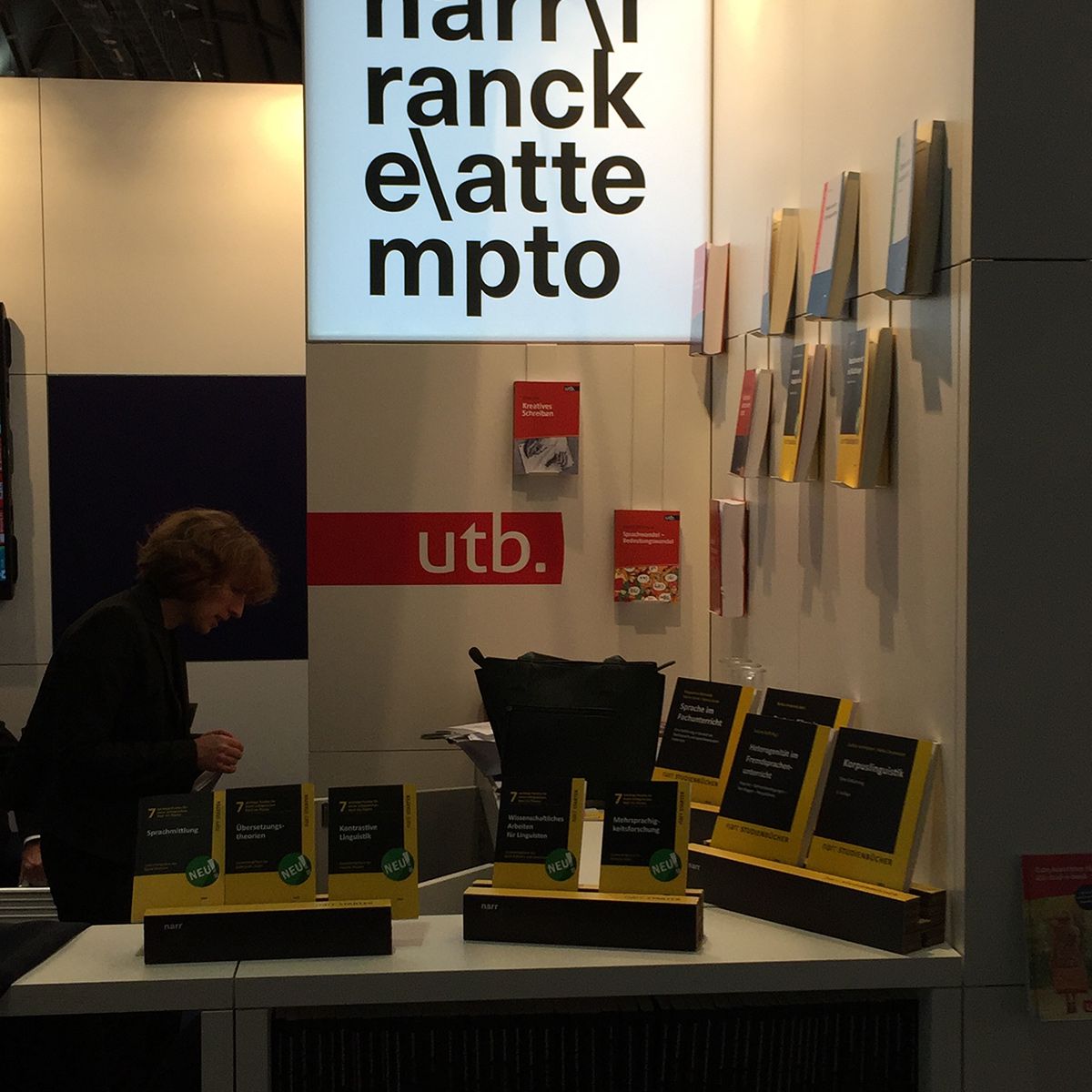 Messestand mit beleuchtetem Schriftzug „Narr Francke Attempto“. Auf dem Tresen liegen schwarze Bücher mit gelbem Rand und grünen Aufklebern. Im Hintergrund sind weitere Bücher und ein rotes „utb.“-Logo sichtbar.