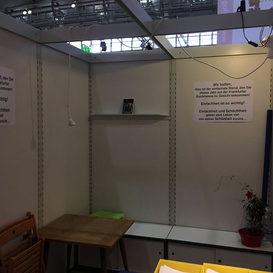 Kleiner Messestand mit weißer Wand, Holztisch, gefalteten Stühlen und roter Topfpflanze. An der Wand hängen mehrere deutschsprachige Schilder mit Zitaten zur Einfachheit und Schönheit.