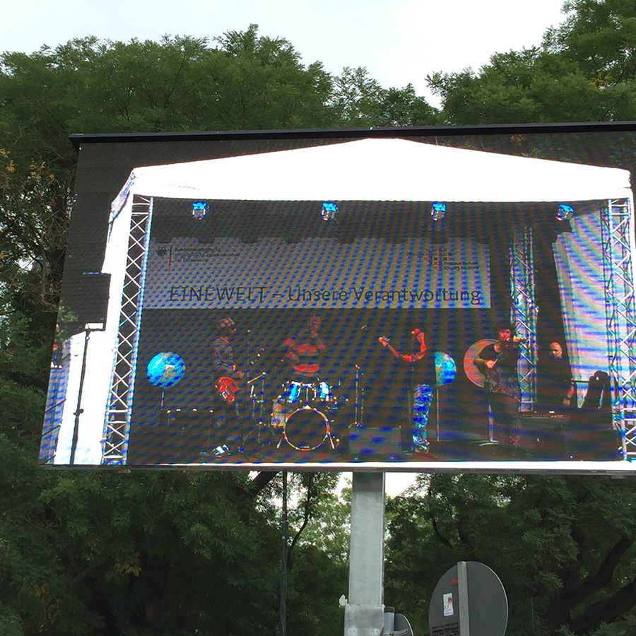 Großbildschirm zeigt eine Live-Musikperformance auf einer überdachten Bühne im Freien. Die Bühne steht in einem baumbestandenen Gelände, Musiker spielen Schlagzeug und Gitarre.