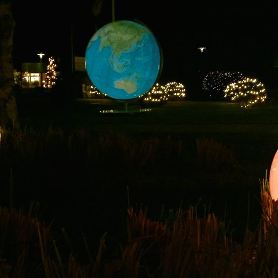 Beleuchtete Globus-Skulpturen bei Nacht, platziert auf einer Rasenfläche mit Büschen und Bäumen. Drei Globen zeigen verschiedene Erdansichten, darunter den Pazifik, Europa und Afrika. Dekorative Lichter im Hintergrund betonen die festliche Atmosphäre.