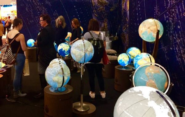 Messestand mit beleuchteten Globen in verschiedenen Größen auf Sockeln. Mehrere Personen betrachten die Ausstellung. Im Hintergrund eine dunkelblaue Wand mit Karten- oder Sternbildmotiv.