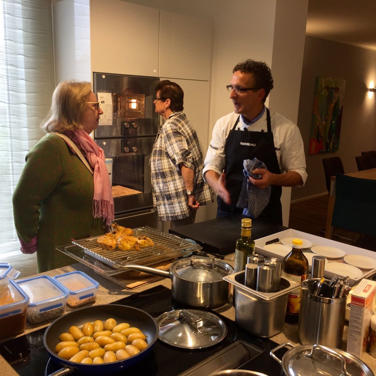 Drei Personen stehen in einer modernen Küche und bereiten Essen zu. Auf dem Herd kochen Kartoffeln, daneben liegen Gebäckstücke auf einem Gitter. Im Hintergrund sind ein Esstisch und ein buntes Gemälde zu sehen.