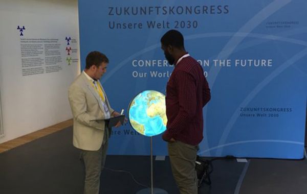 Zwei Personen vor blauem Ausstellungspanel „Unsere Welt 2030“. Einer hält ein Tablet, davor beleuchteter Globus auf Ständer.