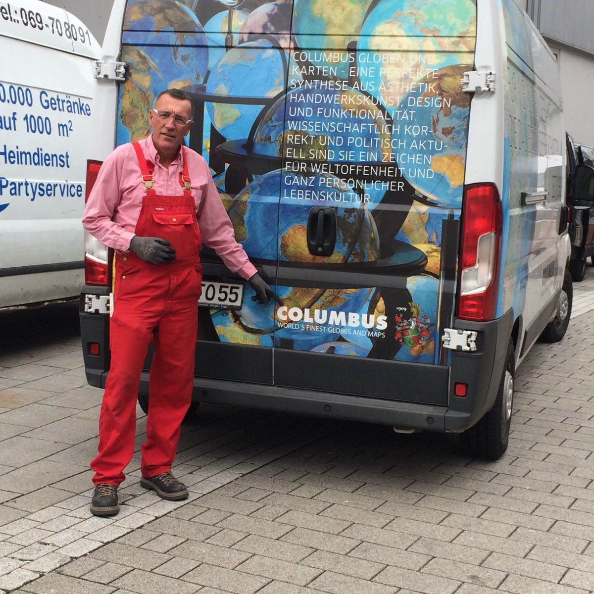 Ein Mann in rotem Overall steht neben einem bunt gestalteten Transporter mit Globusmotiv und dem Schriftzug „COLUMBUS WERKSTATT“.