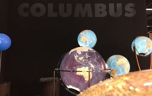 Mehrere Globen vor dunkler Wand mit „COLUMBUS“-Schriftzug. Unterschiedliche Farben und Größen, zentrale Kugel in Violett.