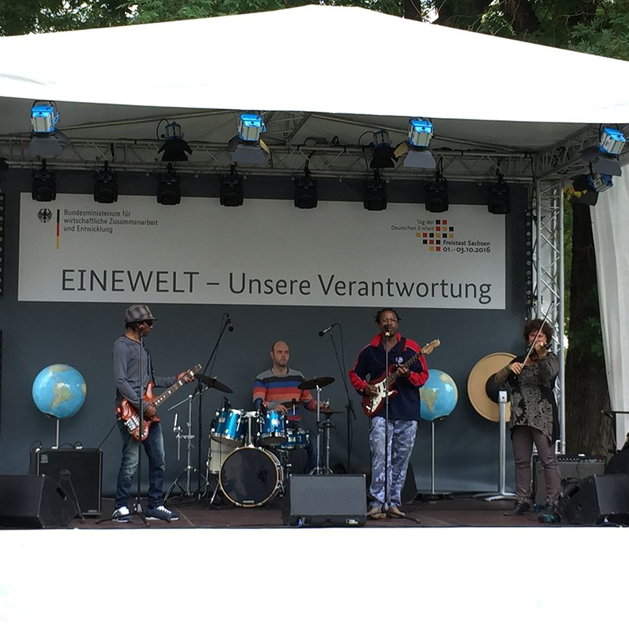 Open-Air-Bühne mit vier Musikern: Gitarre, Bass, Schlagzeug und Geige. Im Hintergrund Banner mit „EINEWELT – Unsere Verantwortung“ sowie Logos deutscher Organisationen. Die Bühne ist mit Globusmodellen dekoriert.
