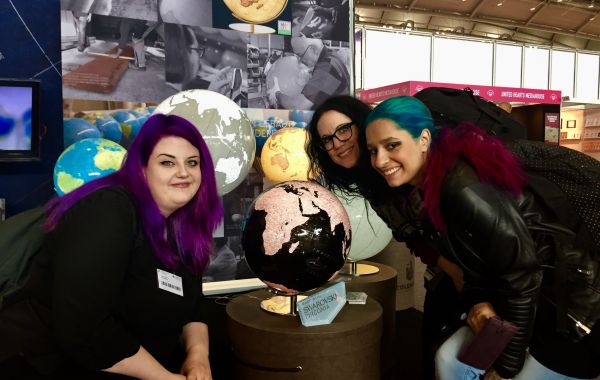 Drei Personen mit auffällig gefärbtem Haar posieren vor beleuchteten Globen auf einem Messestand. Im Hintergrund sind Poster mit Themen wie „DEEP SEA“ und Kartenmotiven sowie weitere Ausstellungsstände sichtbar.