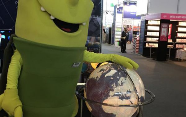 Eine Person in einem grün-gelben Maskottchenkostüm steht neben einem beleuchteten Globus auf Metallständer. Im Hintergrund sind Messestände, Bücherregale und Banner sichtbar.