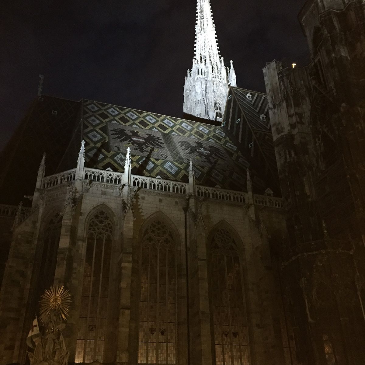 Stephansdom in Wien bei Nacht, mit beleuchtetem Turm und farbigem Ziegeldach. Im Vordergrund Statue und Verkehrsschilder.