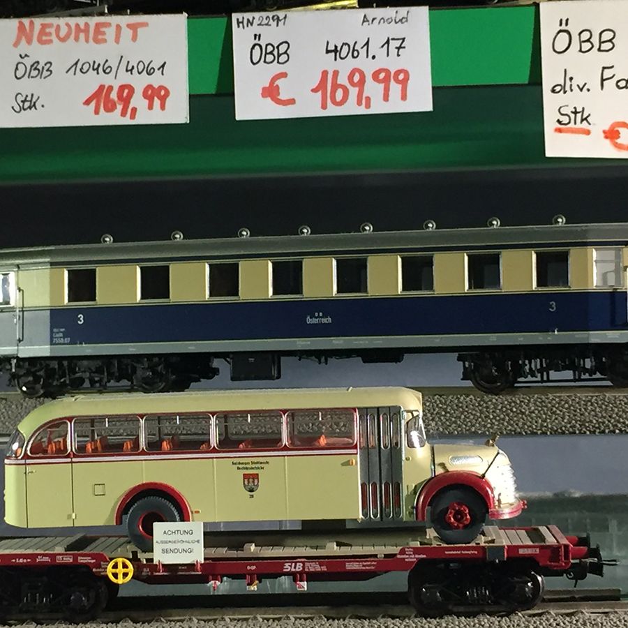 Modellzug „ÖBB 4061.17“ auf Gleis, darunter Modellbus auf Flachwagen. Preisschilder mit Produktinfos im Hintergrund.