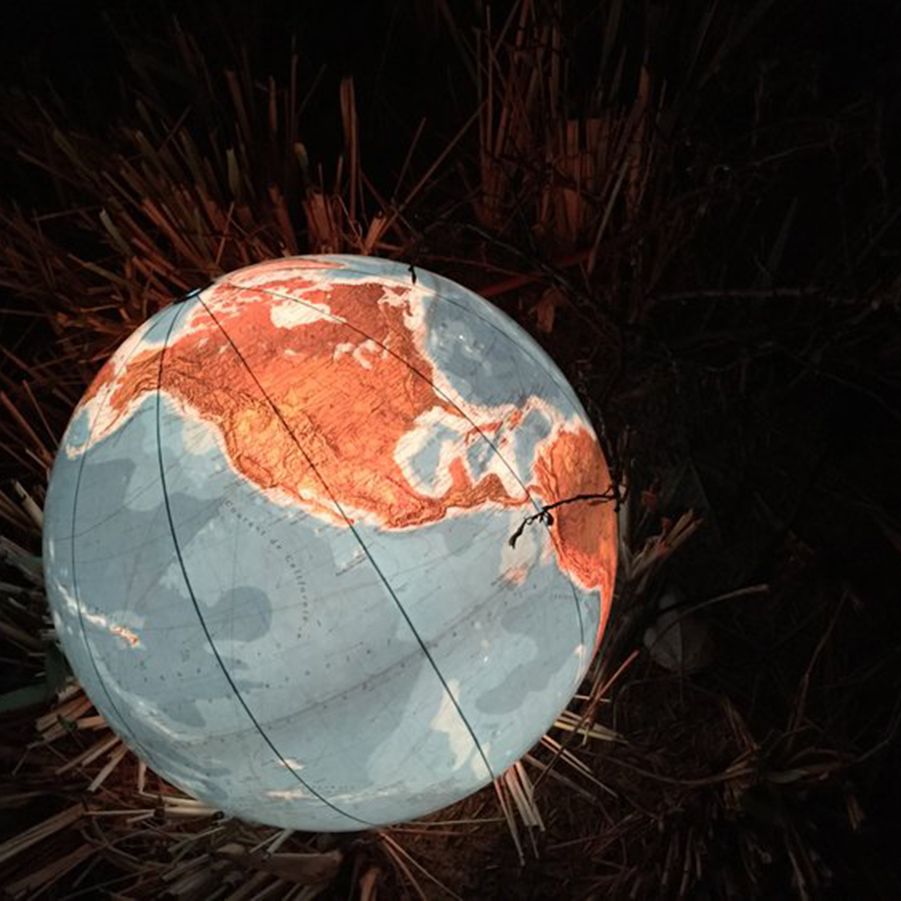 Beleuchteter Globus bei Nacht, platziert zwischen trockenem Gras. Sichtbar sind Nord- und Südamerika sowie Teile des Pazifiks und Atlantiks. Warme Lichttöne heben die Landmassen hervor, kühle Blautöne die Ozeane.
