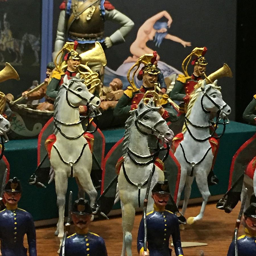 Miniaturfiguren in Paradeformation, darunter berittene Soldaten mit goldenen Helmen und Musiker. Weitere Fußsoldaten in blauen Uniformen, bemalter Hintergrund.