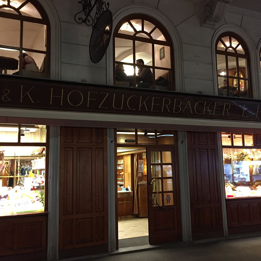 Historisches Gebäude bei Nacht mit beleuchtetem Schaufenster. Schild „K. & K. Hofzuckerbäcker Heiner“, Fenster zeigen Süßwaren. Obergeschoss mit Personen hinter Rundbogenfenstern.