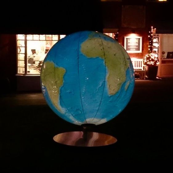 Große beleuchtete Globus-Skulptur im Außenbereich vor einem Gebäude mit Lichterketten. Der Globus zeigt Afrika und Südamerika. Das Gebäude hat große Fenster und warme Innenbeleuchtung. Dunkle Umgebung betont die Lichtwirkung.