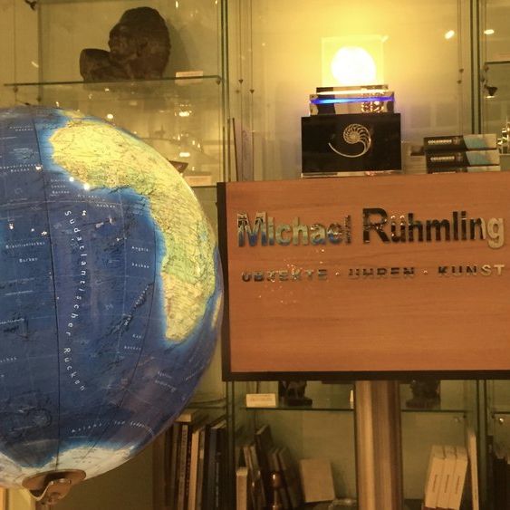 nnenraum mit großem Globus, Holzschild mit der Aufschrift „Michael Ruhmling – OBJEKTE · IDEEN · KUNST“ und beleuchtetem Spiralmotiv darüber. Im Hintergrund Glasregale mit Büsten, Büchern und dekorativen Objekten.