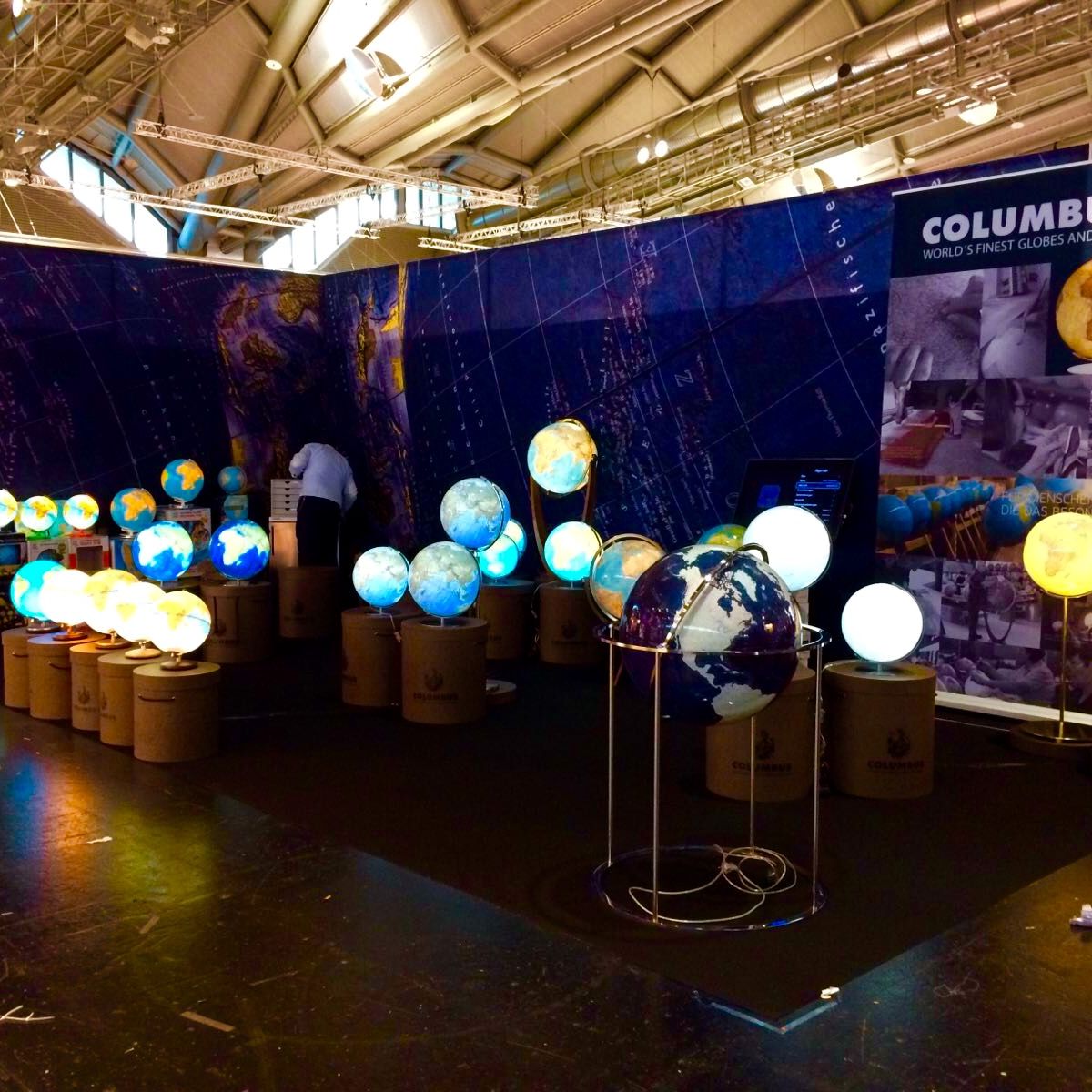Messestand mit beleuchtetem Banner „COLUMBUS – World’s Finest Globes and Maps“. Mehrere Globen in verschiedenen Größen und Designs stehen auf zylindrischen Sockeln. Eine Person betrachtet einen der Globen.