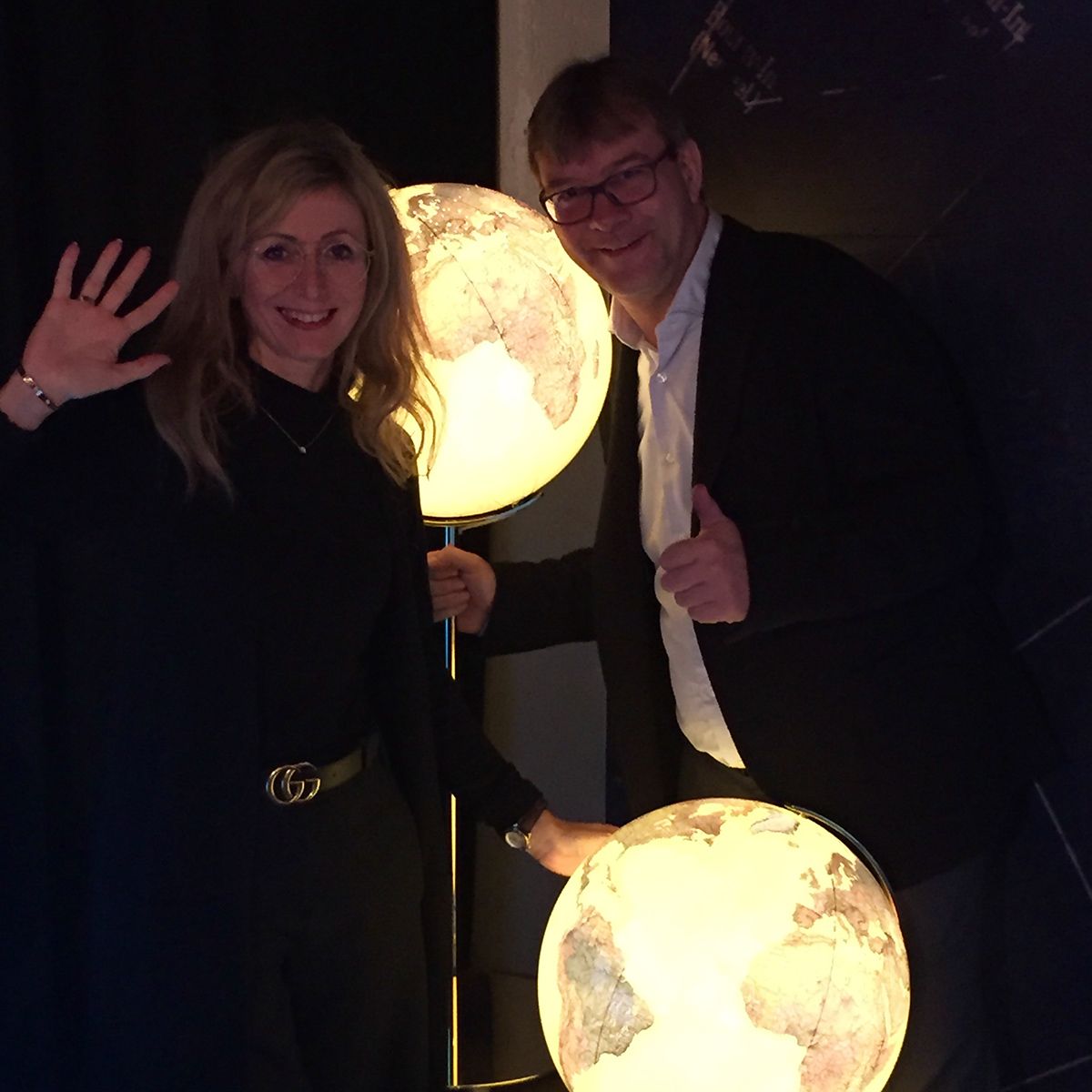 Zwei Personen stehen neben zwei beleuchteten Globen auf Ständern. Eine Person winkt, die andere zeigt einen Daumen hoch. Im Hintergrund sind ein dunkler Vorhang und eine Wand mit kartografischem oder astronomischem Motiv sichtbar.
