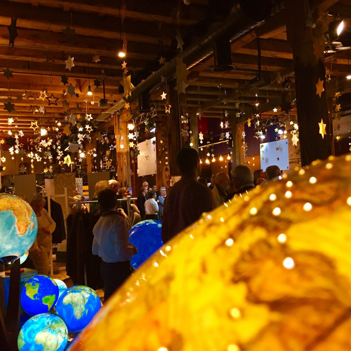 nnenraum mit beleuchteten Globen in verschiedenen Farben und Größen, ergänzt durch hängende Stern-Dekorationen. Mehrere Personen sind anwesend. Die Holzdecke mit sichtbaren Balken und die warme Beleuchtung schaffen eine einladende Atmosphäre.