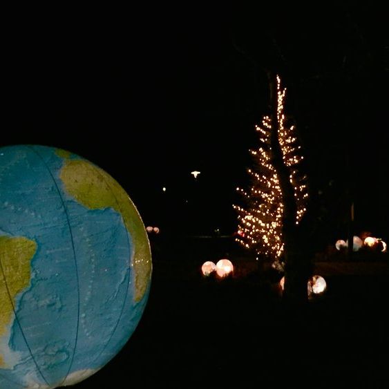 Großes Globusmodell im Vordergrund bei Nacht, zeigt Afrika und Südamerika. Im Hintergrund mehrere leuchtende Kugelobjekte auf dem Boden sowie ein mit Lichterketten geschmückter Baum.