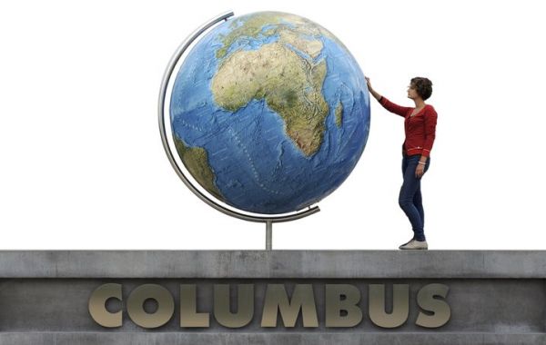 Großer Globus auf einem Sockel mit Fokus auf Afrika, Europa und Asien. Eine Person steht auf einer Plattform mit der Aufschrift „COLUMBUS“ und berührt den Globus.