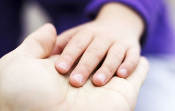 Nahaufnahme von zwei Händen: eine größere Hand, vermutlich eines Erwachsenen, und eine kleinere Kinderhand, die sanft darauf ruht. Das Kind trägt ein violettes Langarmshirt.