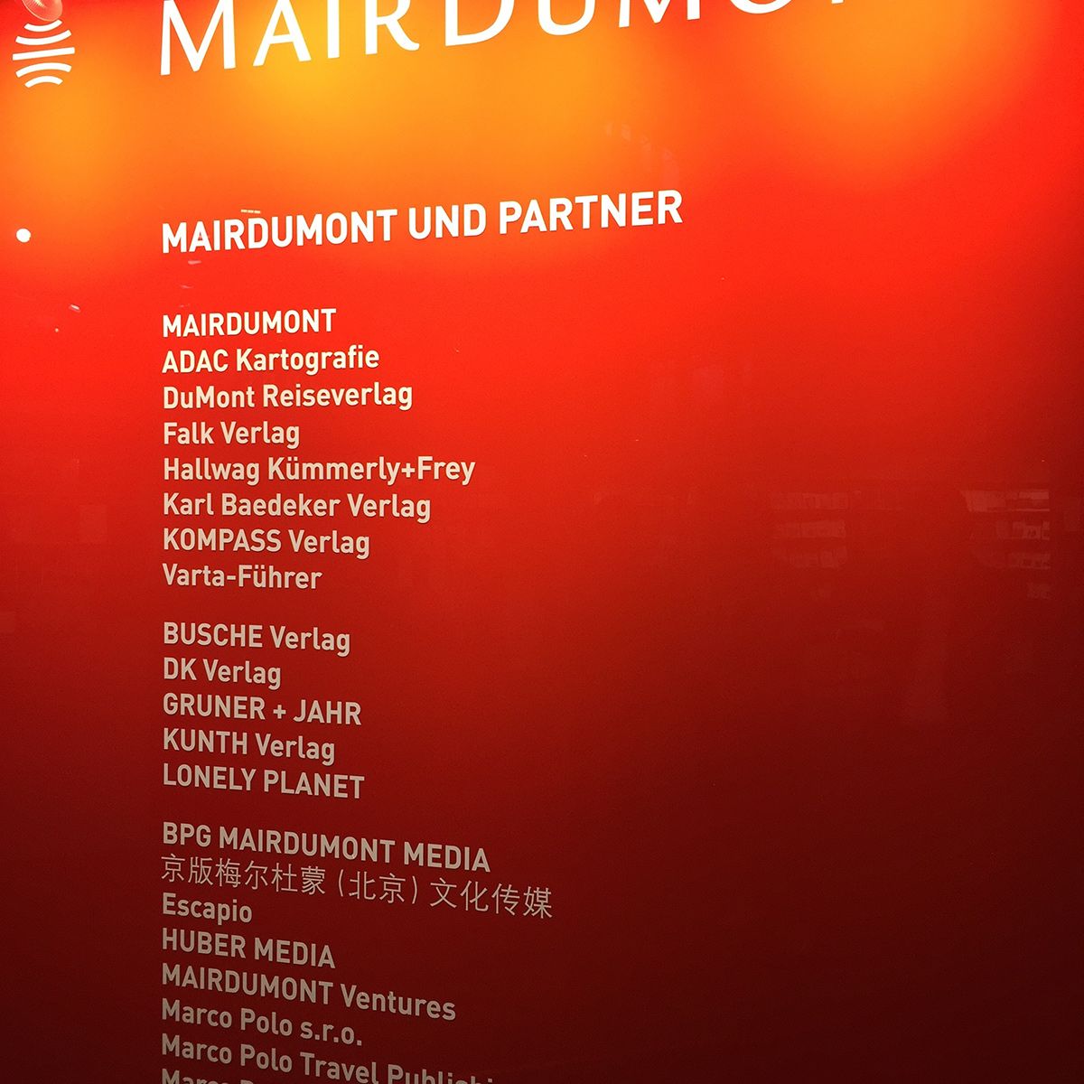 Rote Wand mit weißem Text zu Partnerverlagen von MAIRDUMONT, darunter Namen wie DuMont, Baedeker, Lonely Planet und UTB. Bücher und Logos sind sichtbar.