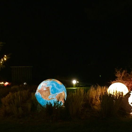 Beleuchtete Globus-Skulpturen bei Nacht, platziert zwischen hohen Gräsern. Mindestens vier Globen zeigen verschiedene Ansichten der Erde, darunter Amerika und Afrika. Links steht ein Baum mit weißen Lichterketten. Weitere Lichtquellen und Dekorationen im