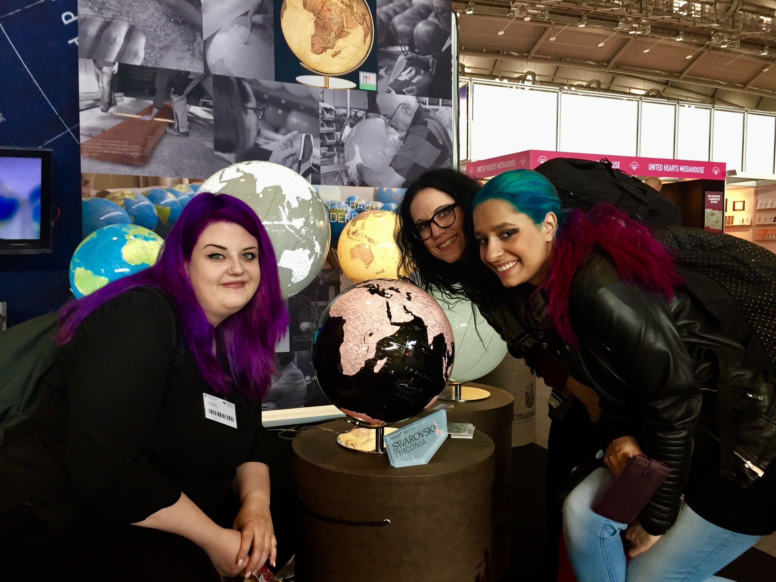 Drei Personen mit auffällig gefärbtem Haar posieren vor beleuchteten Globen auf einem Messestand. Im Hintergrund sind Poster mit Themen wie „DEEP SEA“ und Kartenmotiven sowie weitere Ausstellungsstände sichtbar.