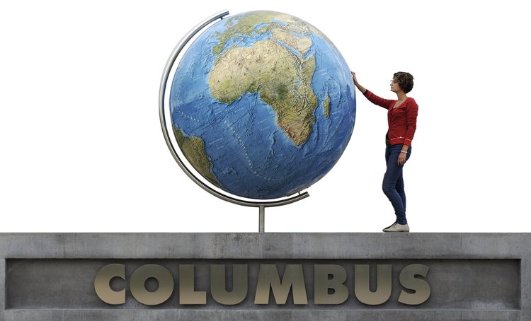 Großer Globus auf einem Sockel mit Fokus auf Afrika, Europa und Asien. Eine Person steht auf einer Plattform mit der Aufschrift „COLUMBUS“ und berührt den Globus.