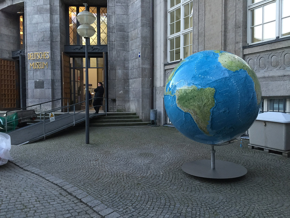 Eingang des Deutschen Museums in München mit großem Globus im Vordergrund. Der Globus zeigt Amerika, das Gebäude hat Glasfassade und goldene Beschriftung.
