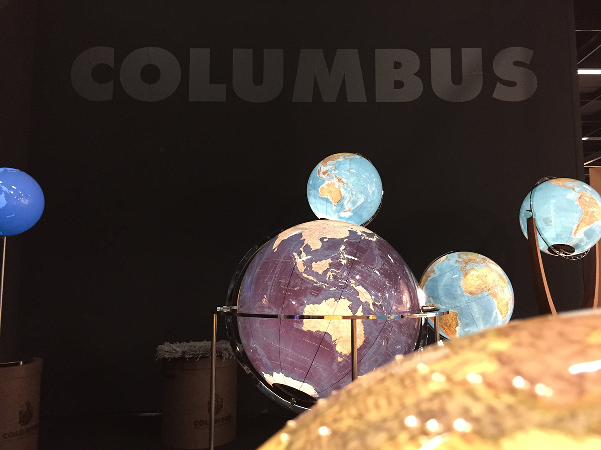 Mehrere Globen vor dunkler Wand mit „COLUMBUS“-Schriftzug. Unterschiedliche Farben und Größen, zentrale Kugel in Violett.
